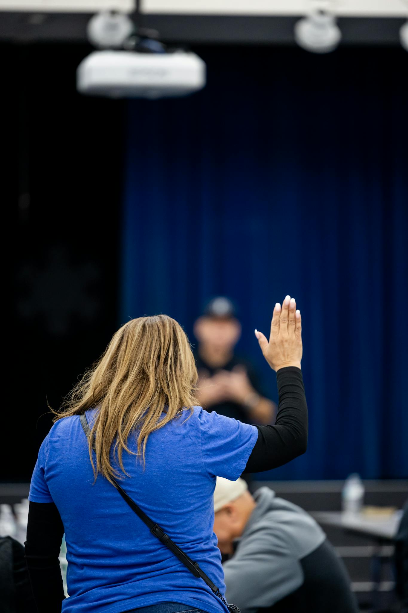 A woman in a blue shirt raises her hand in a classroom or meeting setting, engaging with the speaker.