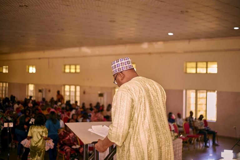 Man in traditional attire speaking to a large audience in a hall.